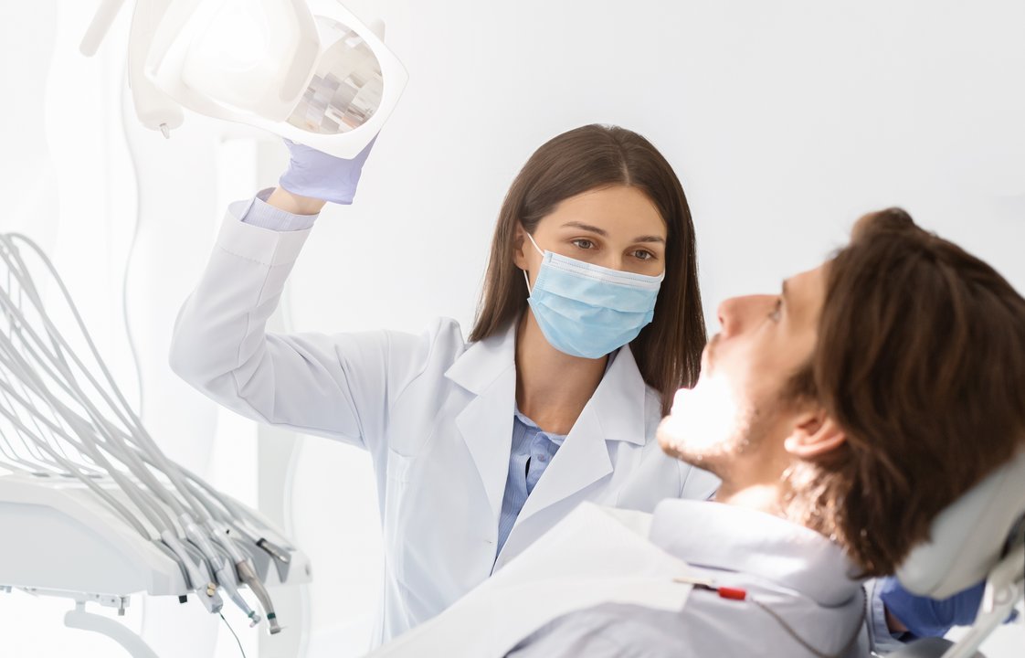 Dentist doctor woman in mask turning on lamp before making check up, patient sitting with open mouth